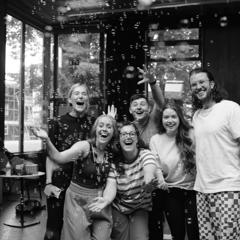 A group of six people joyfully playing with bubbles indoors.